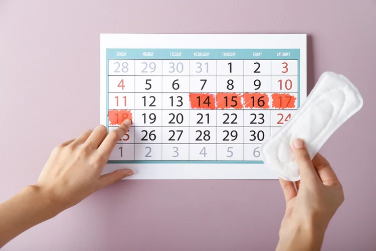 A wall calendar with specific dates marked in red, accompanied by a hand holding a menstrual pad, highlighting menstrual tracking and irregular period management techniques.