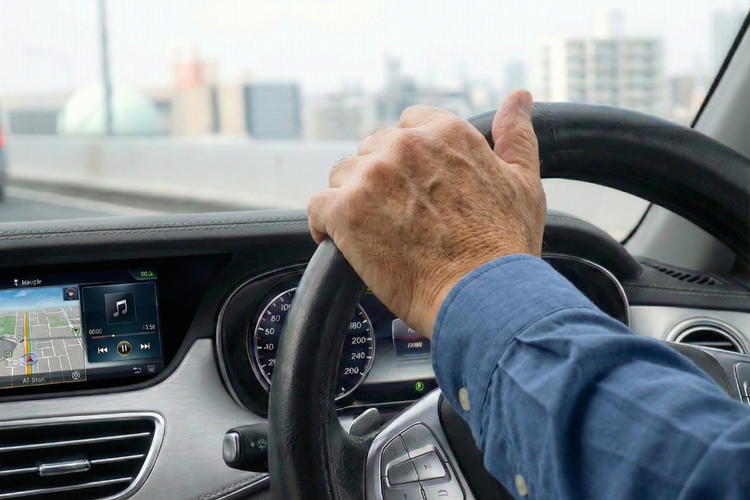 Elderly man driving a car on an expressway with a hand on the steering wheel..