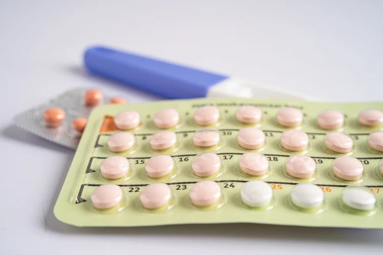 Close-up of a birth control pill pack featuring peach-colored pills in a clear blister strip, alongside a pregnancy test and additional blister packs in the background, symbolizing contraceptive and hormonal therapy options that affect menstrual cycles.