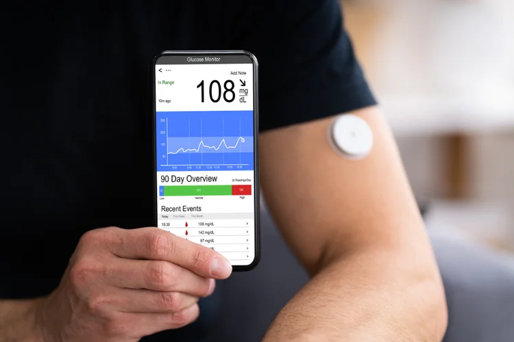 Person wearing a continuous glucose monitoring sensor on arm while checking readings on smartphone.