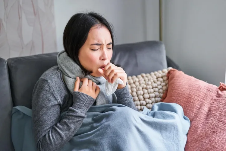 披着围巾、盖着毯子的女性坐在沙发上，手拿纸巾掩口咳嗽